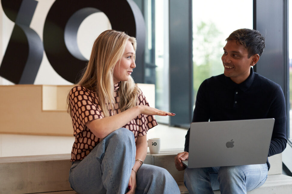 Två personer diskuterar framför en laptop med företagslogotyp i bakgrunden.
